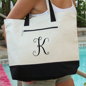 May include: A white canvas tote bag with a black bottom and a black zippered pocket. The bag has a black embroidered letter "K" in a cursive font.