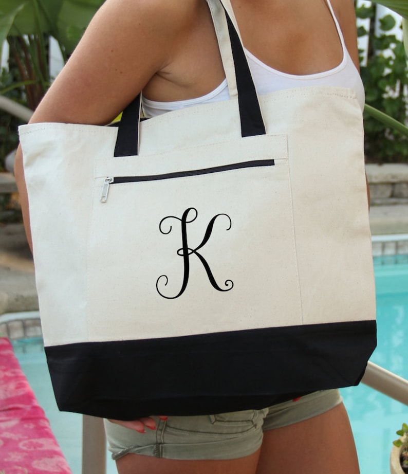 May include: A white canvas tote bag with a black bottom and a black zippered pocket. The bag has a black embroidered letter "K" in a cursive font.