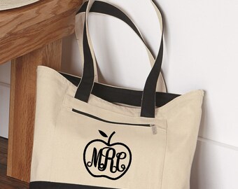 May include: A beige and black canvas tote bag with a zippered pocket. The bag has black straps and a black apple design with the monogram "MRL" on the front.
