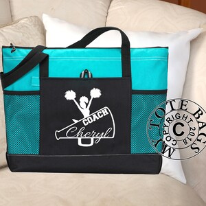 May include: A black and turquoise tote bag with a white design of a cheerleader holding pom-poms and a megaphone with the text "COACH Cheryl".