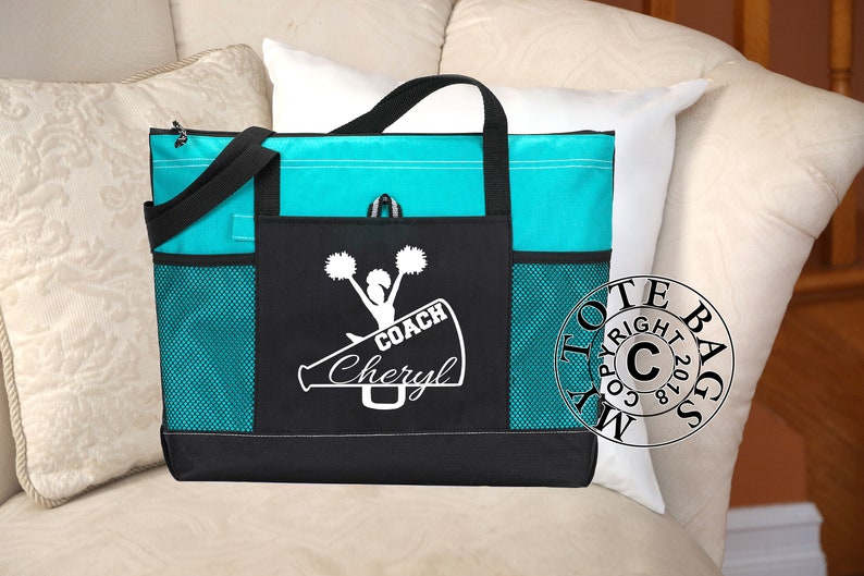 May include: A black and turquoise tote bag with a white design of a cheerleader holding pom-poms and a megaphone with the text "COACH Cheryl".