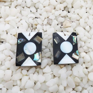 May include: A pair of square-shaped earrings with a black and white geometric design. The earrings are made of mother-of-pearl and have a silver hook.