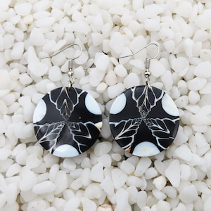 May include: A pair of black and white shell earrings with a floral design. The earrings are round and have a silver hook.