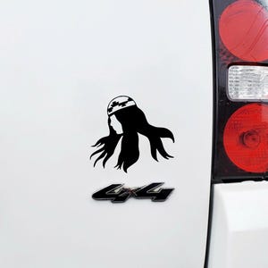 May include: Black and white silhouette of a woman with long hair wearing a patterned head covering. The image is a sticker on the back of a white vehicle.