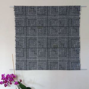May include: Black and white geometric wall hanging with a woven pattern. The hanging is displayed on a white wall with a potted orchid plant in front of it.