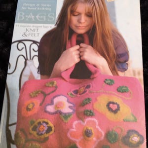 May include: A pink felt bag with floral designs, held by a person. The bag features various colorful flowers in shades of yellow, orange, and purple. The book cover reads "Ella Rae Design & Yarns for hand knitting BAGS book eight 14 exquisite designer bags to KNIT & FELT."