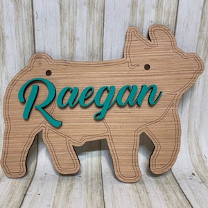 May include: A pig-shaped wooden sign featuring a light brown wood grain finish with dark etched outlines. The name Raegan is displayed in a raised, teal-colored script font across the center of the pig. Two small circular holes are positioned near the top.