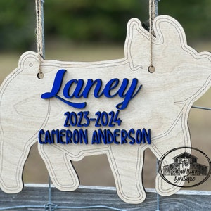May include: A light-colored wooden pig-shaped sign hanging from twine. The sign features the blue text "Laney", "2023-2024", and "CAMERON ANDERSON". A circular logo with the text "The Show Barn Boutique" is visible in the bottom right corner.