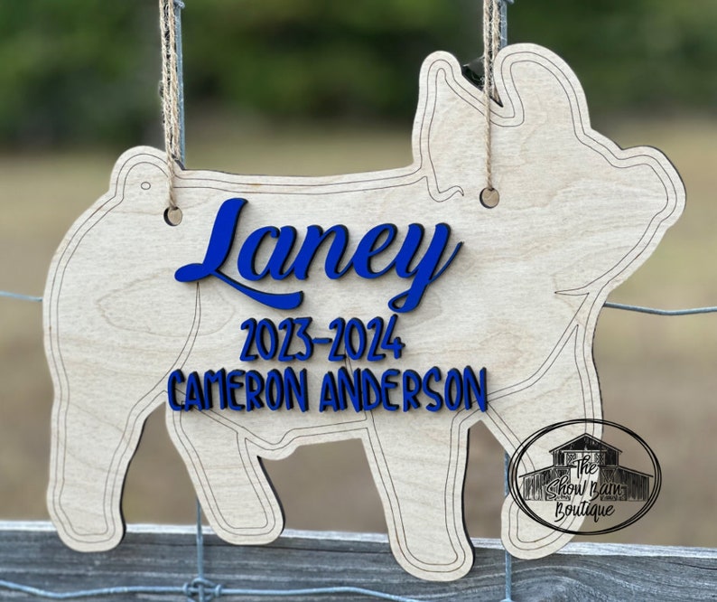 May include: A light-colored wooden pig-shaped sign hanging from twine. The sign features the blue text "Laney", "2023-2024", and "CAMERON ANDERSON". A circular logo with the text "The Show Barn Boutique" is visible in the bottom right corner.