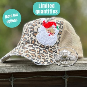 May include: Leopard print trucker hat with a white Santa Claus patch and the text "The Show Barn Boutique" on the back. The hat has a mesh back and a distressed look. The text "Limited quantities" is in a blue circle above the hat and the text "More hat options" is in a blue circle to the left of the hat.