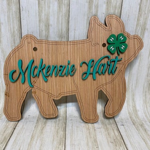 May include: A laser-cut wooden pig sign featuring the name Mckenzie Hart in teal script lettering. The pig shape includes engraved lines and a green four-leaf clover emblem with the letter H in each leaf positioned near the pig's ear area.