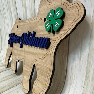 May include: A wooden pig-shaped wall sign featuring a natural wood grain finish with dark engraved outlines. The sign displays the blue script text Kason Philman and a green four-leaf clover emblem with white H letters on each leaf.
