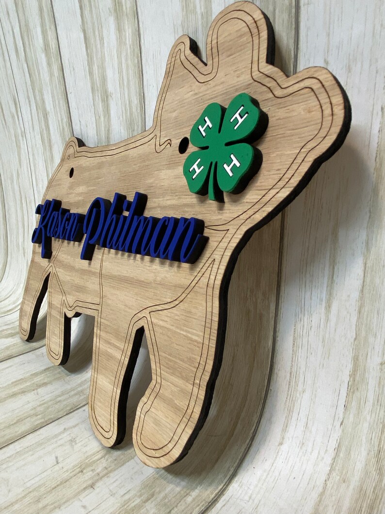 May include: A wooden pig-shaped wall sign featuring a natural wood grain finish with dark engraved outlines. The sign displays the blue script text Kason Philman and a green four-leaf clover emblem with white H letters on each leaf.