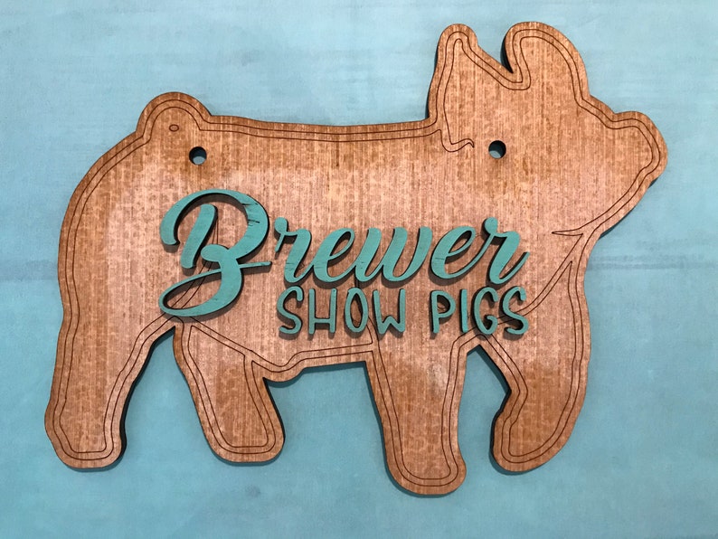 May include: A pig-shaped wooden sign with a natural wood grain finish and engraved outline details. The sign features the teal-colored text "Brewer SHOW PIGS" layered on the front. Two circular holes are positioned at the top for hanging.
