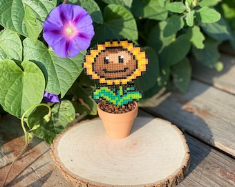May include: A pixel art sunflower character in a terracotta pot, with a smiling face, yellow petals, and green leaves. The pot is sitting on a wooden slice.