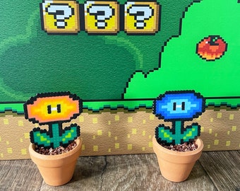 May include: Two potted pixel art flowers. One flower is orange with a yellow centre and the other is blue with a white centre. Both flowers are in terracotta pots and are sitting on a wooden surface.