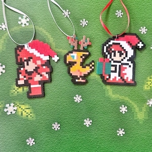 Peut inclure: Trois décorations de Noël représentant des personnages en pixel art. La première décoration est rouge et blanche et représente un personnage portant un bonnet de Père Noël. La deuxième décoration est jaune et représente un personnage ressemblant à un oiseau. La troisième décoration est rouge et blanche et représente un personnage portant un bonnet de Père Noël et tenant un cadeau.