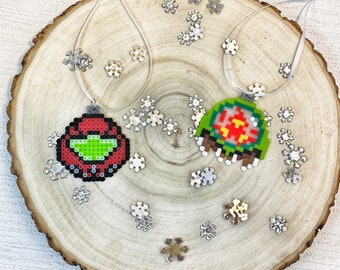 Könnte beinhalten: Zwei Weihnachtsbaumschmuck aus bunten Perlen. Ein Ornament ist rot mit grünen Details und das andere ist grün mit roten Details. Beide Ornamente hängen an einer weißen Schnur auf einem Holzgrund.