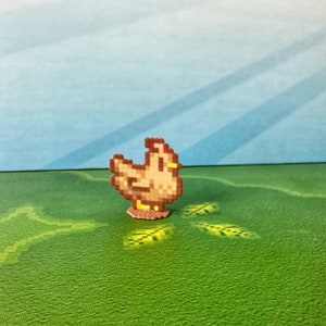 May include: A pixelated chicken figurine in shades of brown, tan, and red, standing on a small, circular base. The chicken is set against a backdrop of a green field and a blue sky, creating a charming, whimsical scene. The design is reminiscent of retro video game graphics.