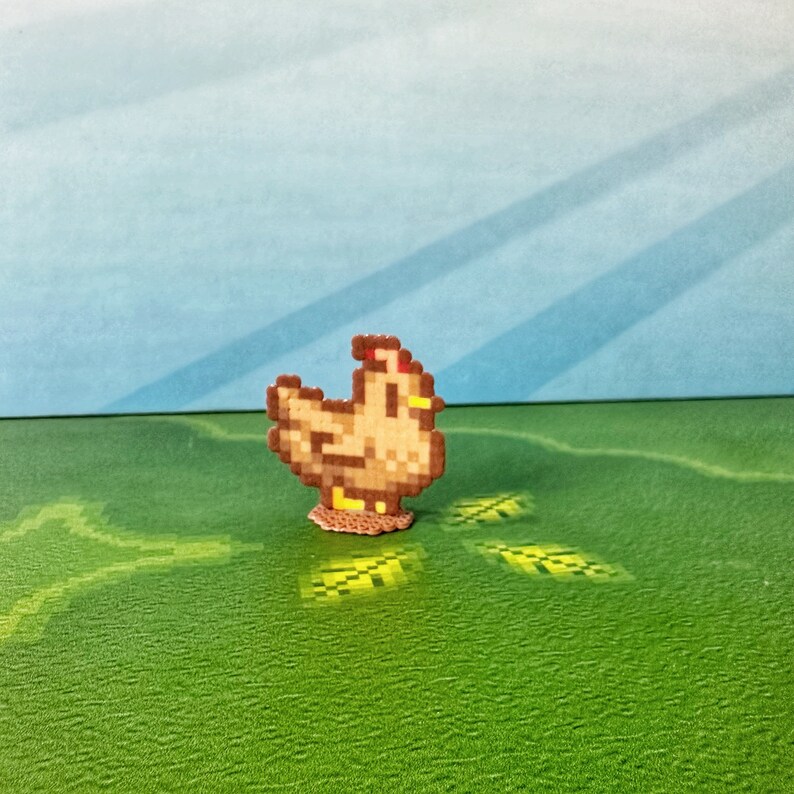 May include: A pixelated chicken figurine in shades of brown, tan, and red, standing on a small, circular base. The chicken is set against a backdrop of a green field and a blue sky, creating a charming, whimsical scene. The design is reminiscent of retro video game graphics.