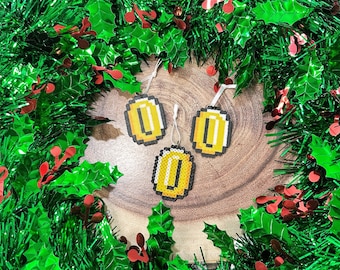 May include: Three gold coin-shaped ornaments with a pixelated design, hanging from white string on a green and red garland.