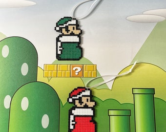 Puede incluir: Dos adornos navideños de pixel art con personajes de Super Mario. El primer adorno es verde y presenta a un Mario vestido de verde con un bloque de signo de interrogación. El segundo adorno es rojo y presenta a un Mario vestido de rojo con un bloque de signo de interrogación.