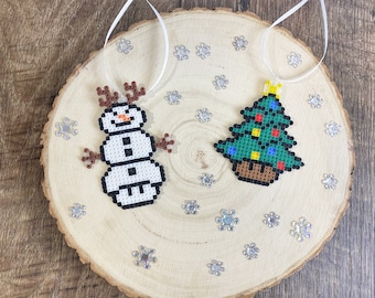 May include: Two Christmas ornaments made of colorful beads. One ornament is a snowman with brown antlers and a black top hat. The other ornament is a green Christmas tree with red, yellow, and blue ornaments.