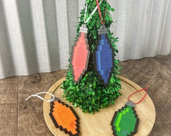 May include: Four pixelated Christmas ornaments, two red, one blue, and one green, hanging on a small artificial Christmas tree. The ornaments are on a wooden surface.