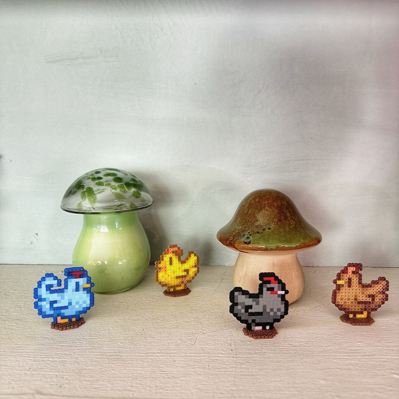 May include: A collection of decorative items featuring two ceramic mushroom sculptures and four pixelated chicken figurines. The mushrooms have green and brown tones, while the chickens are blue, yellow, gray, and brown. The items are arranged on a white surface.
