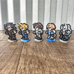 Op de afbeelding: Vijf pixel art figuren van personages uit een videogame. De figuren zijn gemaakt van gekleurde kralen en staan op kleine blauwe bases. De personages dragen allemaal verschillende outfits en hebben verschillende kapsels.