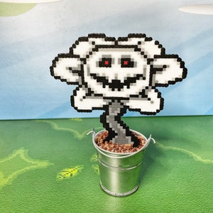May include: A white and gray pixel art flower with red eyes and a toothy grin, sitting in a silver metal bucket filled with brown beads.