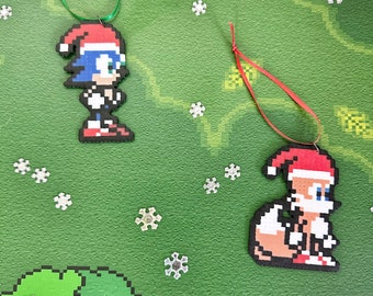 May include: Two Christmas ornaments featuring pixelated characters from the Sonic the Hedgehog video game series. The ornaments are red and white with blue and yellow accents. The first ornament features Sonic the Hedgehog wearing a Santa hat. The second ornament features Tails the Fox wearing a Santa hat.