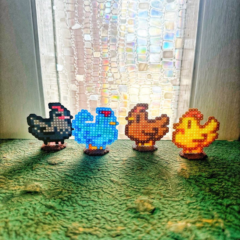 May include: Four pixel art chicken figurines in black, blue, brown, and yellow. Each chicken has a unique design and stands on a small base. The figurines are arranged on a green surface in front of a window.
