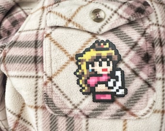 May include: A pixelated character with blonde hair and a pink dress is sewn onto a brown and white plaid fabric. The character is wearing a crown and holding a white object.