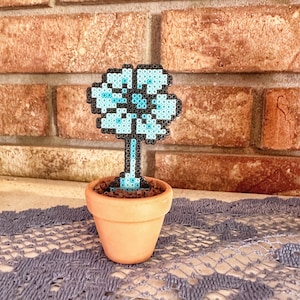 May include: A blue and black pixel art flower in a terracotta pot. The flower is made of small beads and is sitting on a gray lace tablecloth.