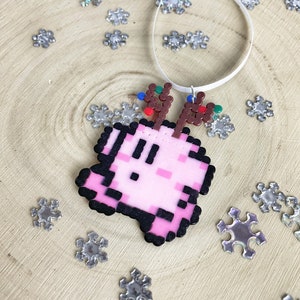 May include: Pink pixelated reindeer ornament with brown antlers and colorful lights. The ornament is hanging from a white ribbon on a wooden background with silver snowflakes.