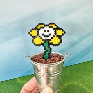 May include: A yellow flower with a smiling face made of perler beads in a small silver bucket with brown soil.