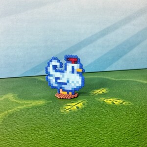May include: A pixelated chicken figurine in shades of blue, white, red, and yellow. The chicken is standing on a brown base, with a green and blue background. The figurine is a charming, handcrafted item.