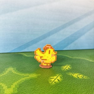 May include: A pixelated yellow chick pin with a brown base. The chick has a small pink accent on its head. The pin is set against a green and blue background.