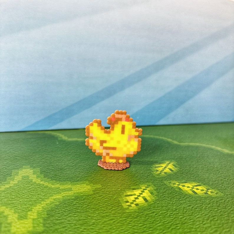 May include: A pixelated yellow chick pin with a brown base. The chick has a small pink accent on its head. The pin is set against a green and blue background.