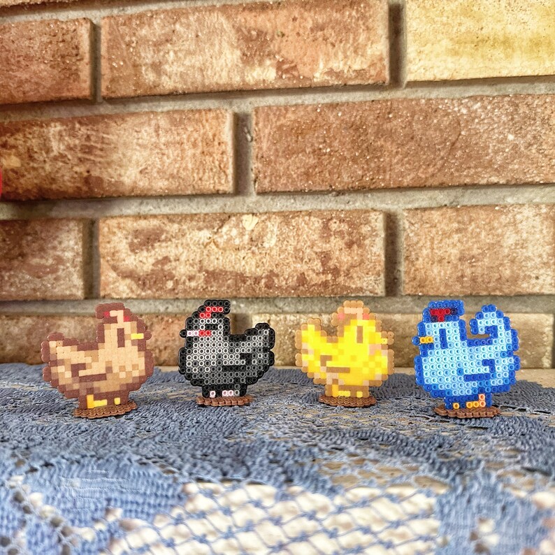 May include: Four pixel art chickens in brown, black, yellow, and blue. Each chicken is made of small, colorful beads and stands on a brown base. The chickens are arranged in a row against a brick wall.