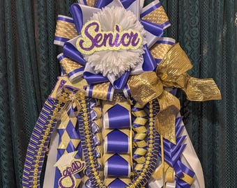 May include: A large homecoming mum decoration featuring layers of royal blue, gold, and white ribbons. The top includes a white flower with a purple glitter cutout reading Senior, while a smaller purple glitter cutout reads grad with a cap.