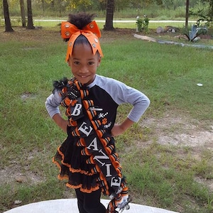 May include: A large, decorative homecoming mum accessory featuring black and orange ribbons, silver glitter lettering spelling BEANIE, and small paw print accents. The accessory is worn over a black and gray raglan shirt and a black tutu skirt.