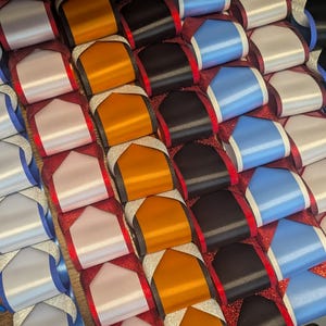 May include: Close-up of a decorative craft with rows of colorful satin ribbons. The ribbons are arranged in a grid pattern, with colors including white, orange, black, and blue. The base is a glittery red material.