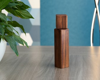 May include: A wooden bottle stopper with a dark brown finish. The stopper is made of two pieces, a square top and a rectangular base.