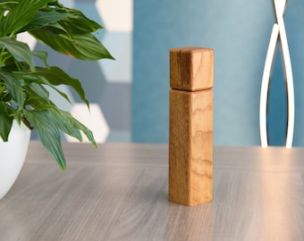 Könnte beinhalten: Ein Salz- und Pfefferstreuer aus Holz mit quadratischer Oberseite und zylindrischem Körper. Der Streuer ist aus hellbraunem Holz mit natürlicher Maserung.