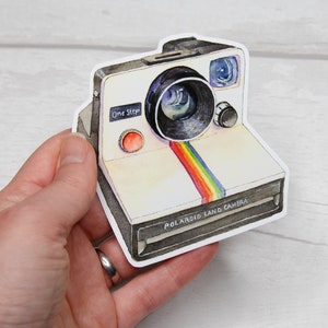 Pu&ograve; includere: Una Polaroid Land Camera bianca con una striscia arcobaleno sul davanti. La fotocamera ha un obiettivo nero e un pulsante rosso con la scritta "One Step".