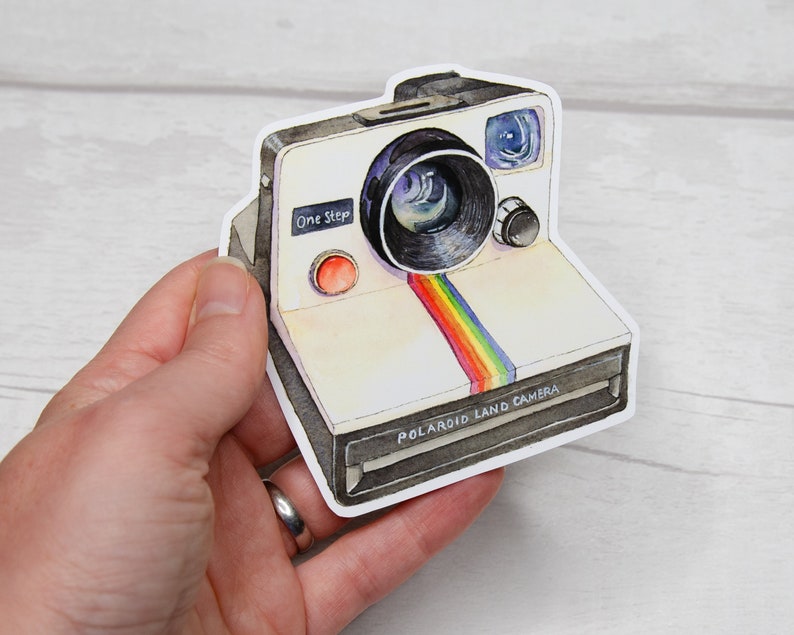 Pu&ograve; includere: Una Polaroid Land Camera bianca con una striscia arcobaleno sul davanti. La fotocamera ha un obiettivo nero e un pulsante rosso con la scritta "One Step".