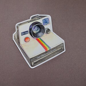 Pu&ograve; includere: Un adesivo di una Polaroid Land Camera in stile vintage con una striscia arcobaleno e il testo "One Step" e "Polaroid Land Camera".