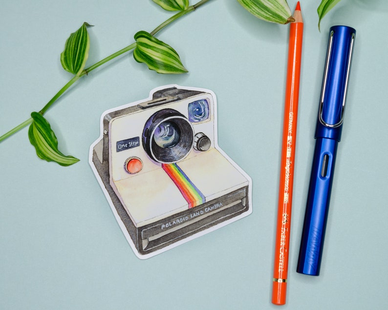 Pu&ograve; includere: Un adesivo con una Polaroid Land Camera in stile vintage con corpo bianco, strisce arcobaleno e il testo "One Step" e "Polaroid Land Camera".